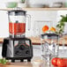 A commercial blender with a red liquid in it and a tomato on top.