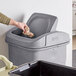 A hand throwing a paper into a Toter gray square swing door lid on a trash can.