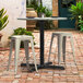 A Lancaster Table and Seating black outdoor table base with a table and two stools on a brick patio.