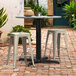 A Lancaster Table & Seating Excalibur outdoor table with a black base on a brick patio with white stools.