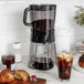 An OXO cold brew coffee maker on a table with a glass of iced coffee and a croissant.