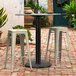 A Lancaster Table & Seating Excalibur black outdoor table base with two white bar stools on a brick patio.