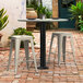 A black Lancaster Table & Seating Excalibur bolt down table base with stools on a brick patio.