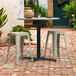 A Lancaster Table & Seating black outdoor table with two stools on a brick patio.