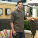 A man wearing an olive short sleeve chef coat with a chair on a patio.