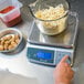 A person weighing noodles in a bowl on a San Jamar digital portion scale.
