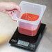 A hand using a San Jamar black kitchen scale to weigh a container of red sauce.