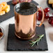 A copper mug with ice and liquid on a black Acopa slate coaster with food on it.