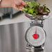 A hand using a San Jamar portion scale to weigh a bowl of salad on a counter.