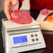 A hand weighing a piece of raw meat on a San Jamar digital portion scale.