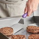 A person using a Vollrath Jacob's Pride stainless steel hamburger turner with a purple handle to cook burgers on a pan.