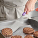A person using a Vollrath Jacob's Pride Hamburger Turner with a purple handle to cook burgers.