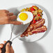 A person using an OXO stainless steel spatula to serve bacon and eggs on a plate.