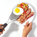 A hand using an OXO stainless steel spatula to serve a fried egg on a plate of bacon and eggs.