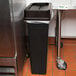 A black rectangular Continental wall hugger trash can on a counter with a plastic bag over it.