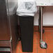 A black rectangular Continental wall hugger trash can with a black handle sitting on a counter.