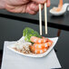A person holding chopsticks over a plate of shrimp and broccoli on an Arcoroc square porcelain plate.