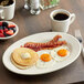 An Acopa ivory oval platter with a plate of breakfast food including pancakes and bacon on a table.