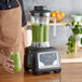 A person in a brown apron holding an AvaMix commercial blender with a green smoothie inside.