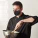 A man wearing a Mercer Culinary black face mask mixing food in a bowl.