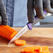 A person in black gloves using a Dexter-Russell purple-handled paring knife to cut a carrot on a wooden cutting board.