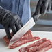 A person wearing black gloves uses a Dexter-Russell Santoku knife to cut a piece of meat.