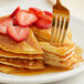A fork on a plate with a stack of Golden Dipt pancakes topped with strawberries and syrup.