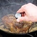 A hand using a Taylor digital pocket probe thermometer to check the temperature of cooked meat in a pan.