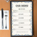An 8 1/2" x 14" menu with Mediterranean border on a wood table with a fork and knife.