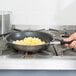 A hand using a Vollrath stainless steel frying pan with a black TriVent handle to cook scrambled eggs on a stove.