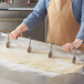 A person using an Ateco stainless steel rod to cut dough on a table in a professional kitchen.