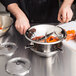 A person using a Tellier stainless steel food mill to prepare carrots in a metal bowl.