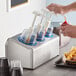 A person pouring ketchup into a Steril-Sil stainless steel condiment dispenser with blue pump lids.