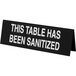 A black Cal-Mil table tent sign with white text that reads "This table has been sanitized"