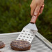 A hand using an Outset stainless steel slotted turner to flip burgers on a grill.