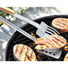 Stainless steel tongs with bamboo handles gripping seasoned meat on a grill.