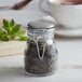 A Tablecraft glass condiment jar with a stainless steel lid filled with green tea leaves.
