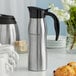 A silver and black Choice stainless steel coffee carafe with a coffee cup on a table.