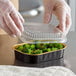 A hand in plastic gloves holds a ChoiceHD black and gold mini foil container with broccoli inside.