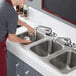 A person using a Regency stainless steel three compartment drop-in sink to wash dishes.