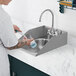 A person washing dishes in a Waterloo stainless steel drop-in sink with a gooseneck faucet.