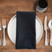 A black Milan satin band cloth napkin folded on a plate with silverware on a table.
