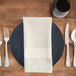 A place setting with a cream Milan satin band Snap Drape cloth napkin and silverware on a black plate.