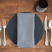 A blue Milan birdseye Snap Drape cloth napkin on a plate with silverware on a table.