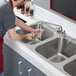 A man using a 20 gauge stainless steel Regency three compartment sink with a faucet to wash a glass.