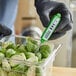 A hand in black gloves holding a green AvaTemp digital pocket probe thermometer in a container of lettuce.