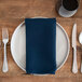 A plate with a Snap Drape navy birdseye cloth napkin and silverware on a wooden table.