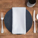 A white Milan birdseye cloth napkin on a wooden table with a plate, fork, and knife.