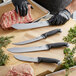 A person wearing black gloves holds a Schraf Butcher Knife with a black TPR grip handle and cuts a piece of meat.