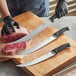 A person in gloves using a Schraf Cimeter knife to cut raw meat on a cutting board.